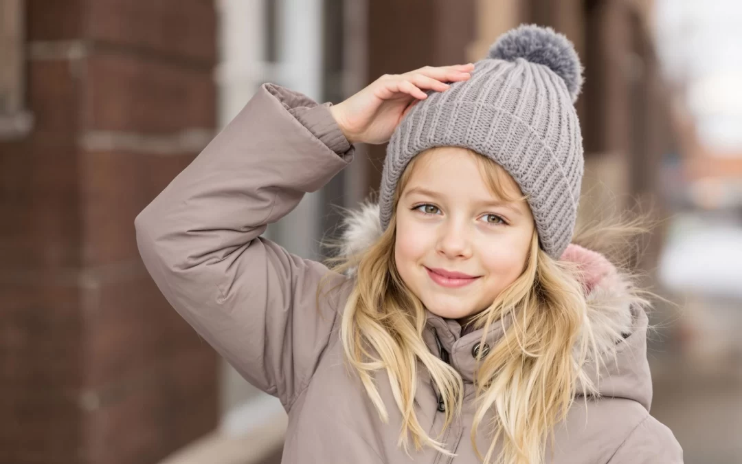Girl wearing a cozy winter coat and gray knitted hat, smiling outdoors, representing winter conditions and children's activities relevant to head lice transmission.